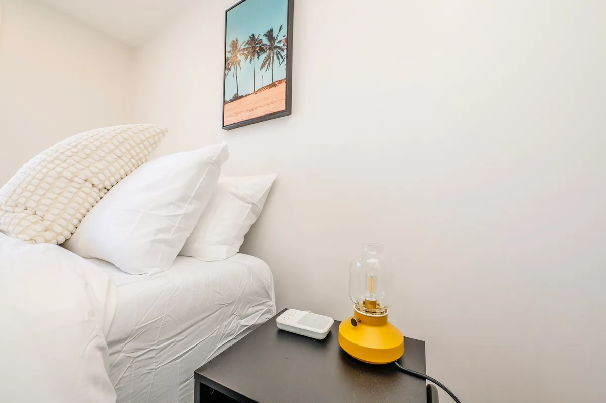 A cozy bedroom featuring large white pillows on a neatly made bed, bathed in soft sunlight streaming through a nearby window. A yellow lamp casts a gentle glow, while a framed beach photograph adds a touch of relaxation and escape.