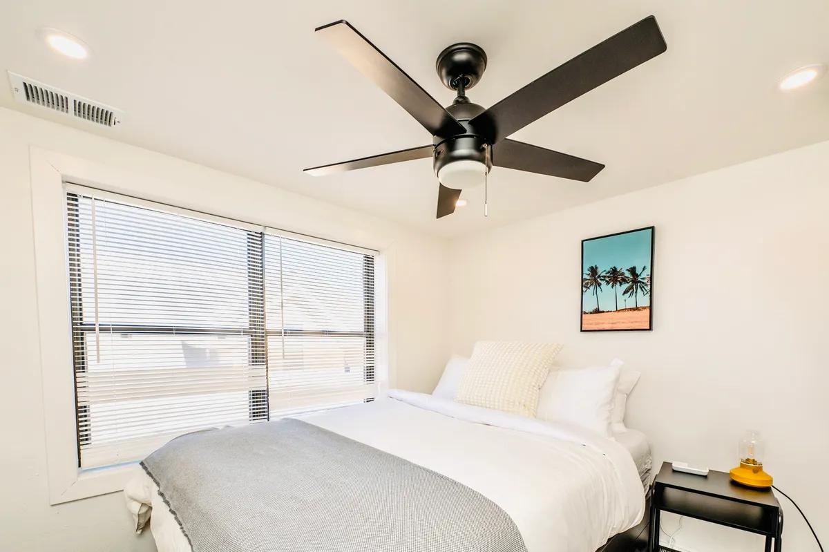 A spacious bedroom with a king-sized bed, ceiling fan, palm tree artwork, and large windows that flood the room with natural light. The peaceful setting makes it perfect for unwinding and enjoying restorative downtime.