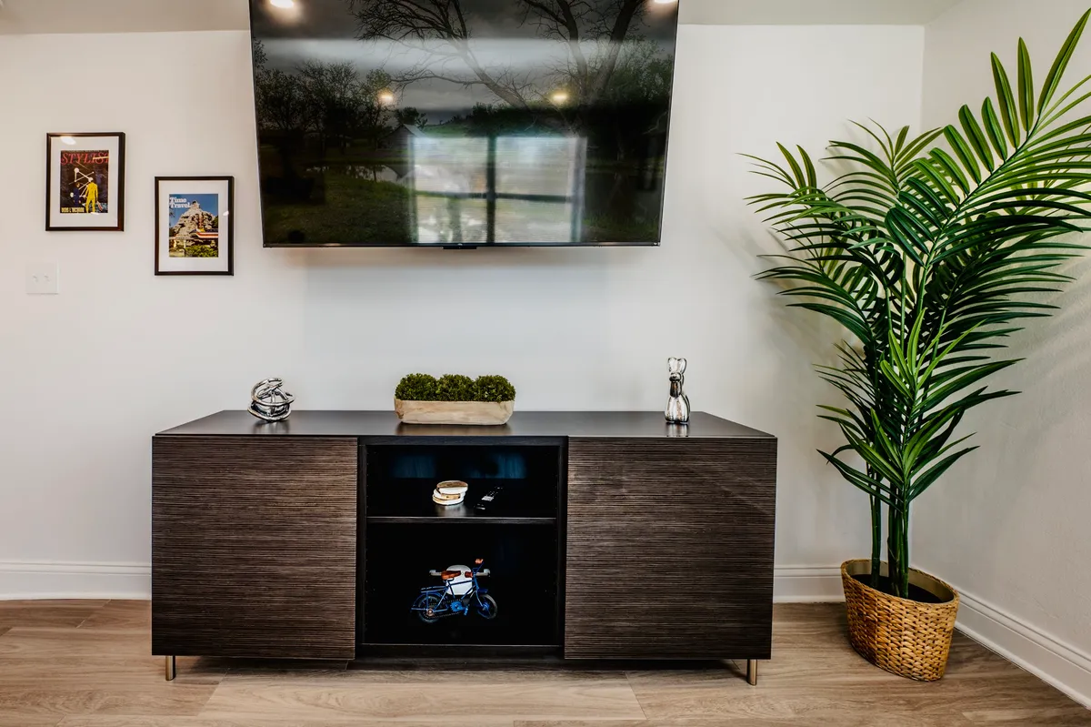 A modern, inviting living area with a television, decorative shelving, houseplants, and framed pictures that suggest a connection to local culture and personal expression. The space feels both comfortable and lively, ideal for winding down after a day out.