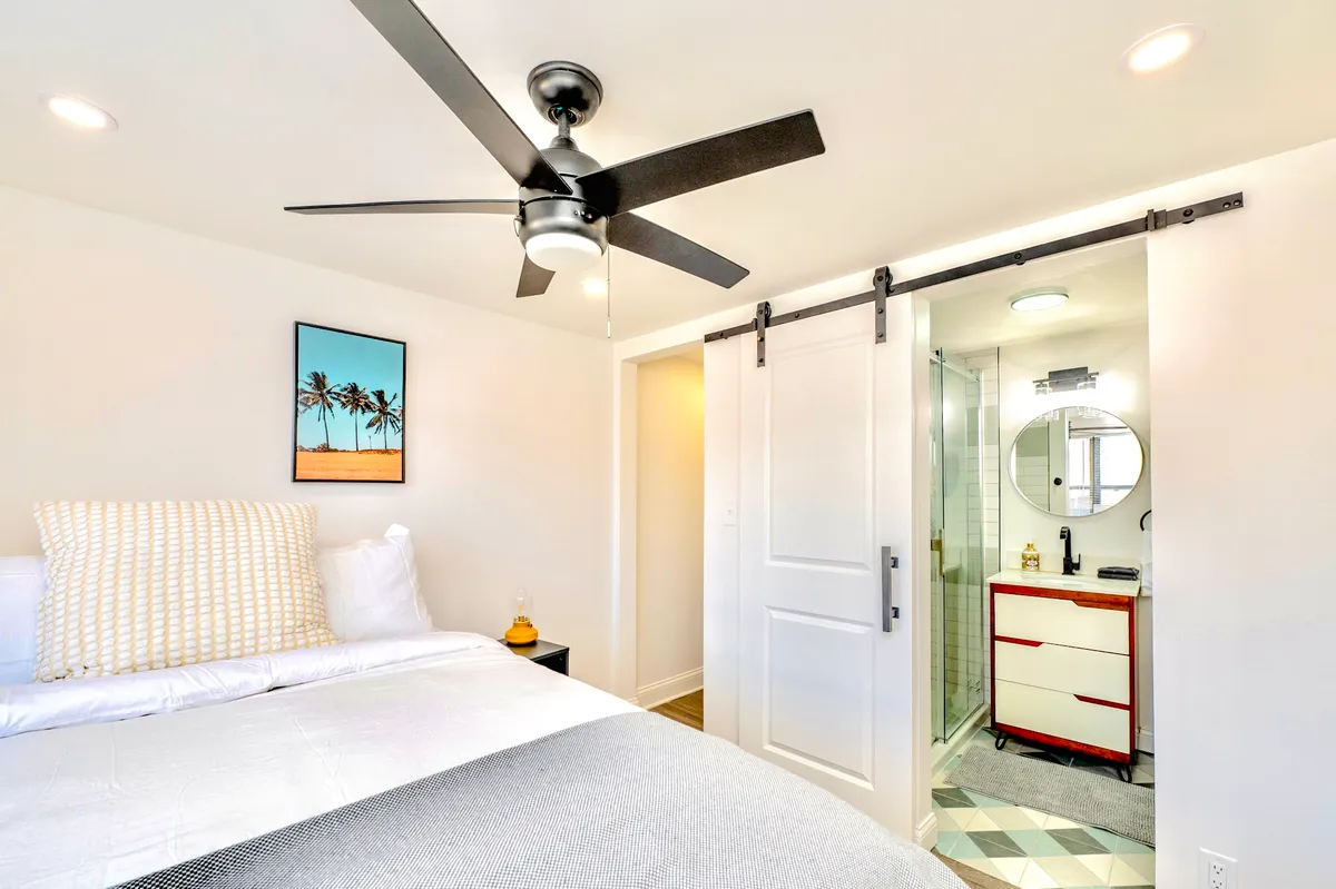A serene bedroom featuring a plush bed adorned with decorative pillows, framed palm tree artwork above the headboard, and a modern ceiling fan. A sliding door leads to the bathroom, and sunlight gently filters in, creating a peaceful atmosphere ideal for relaxation.