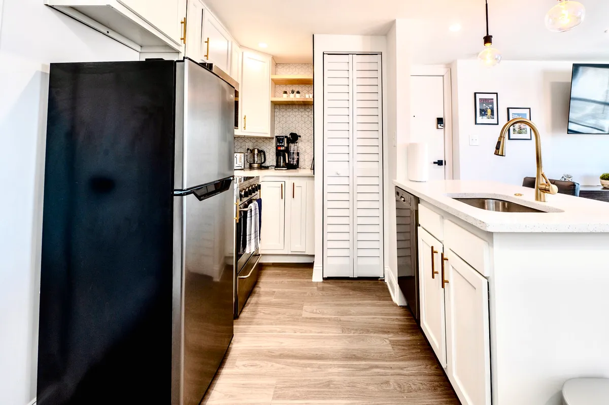 This image features a contemporary kitchen with a striking black refrigerator, light wooden flooring, white cabinetry with gold fixtures, and a spacious island with an integrated sink. The open design suggests a space perfect for hosting, experimenting with recipes, or enjoying a morning coffee.