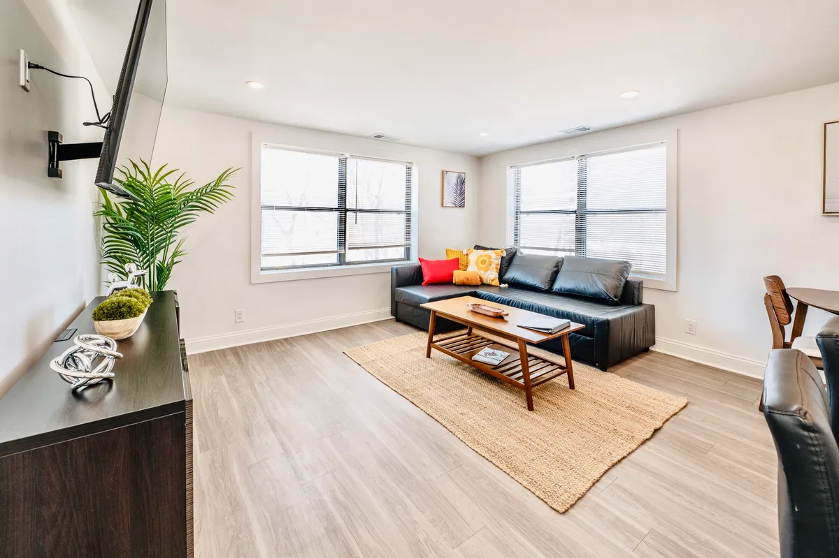 A bright and airy living room featuring a soft sofa, a sleek coffee table, a television, large windows letting in daylight, and a lush indoor plant. The arrangement promotes both socializing and quiet downtime.