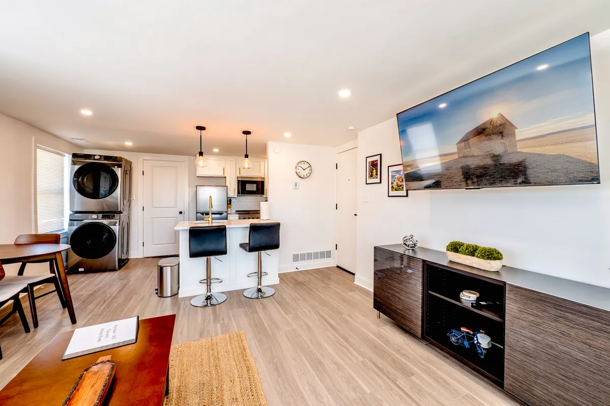 This image displays a cozy kitchen area complete with bar stools, a dining table, and a TV showing a landscape image. The open layout connects the cooking and dining spaces, inviting laughter over meals or relaxed evenings watching TV after dinner.