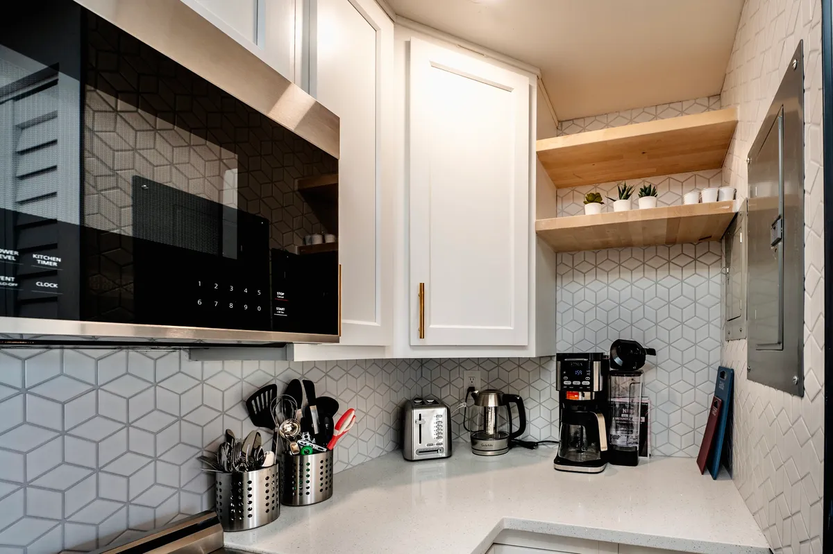 A cozy and functional kitchen area featuring a microwave, coffee maker, toaster, and various kitchen utensils, with touches of greenery from plants on a shelf. The setup is practical yet inviting, emphasizing the convenience of preparing meals or brewing coffee at home.