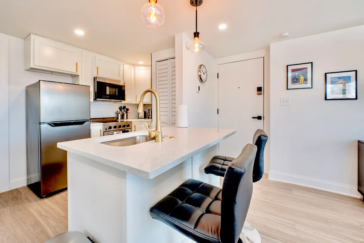 This image showcases a modern kitchen with a central island, gold faucet, and stylish bar stools. High-end appliances are visible, and tasteful artwork adorns the wall, creating a welcoming space for cooking and entertaining. The presence of bar stools highlights opportunities for casual dining or morning coffee gatherings.