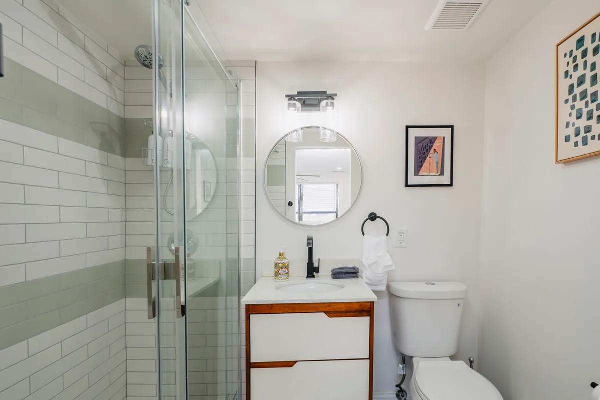 A modern bathroom featuring a glass shower enclosure, round mirror, vanity with wooden accents, and decorative artwork. The inviting setting is ideal for refreshing morning routines or unwinding after a busy day.
