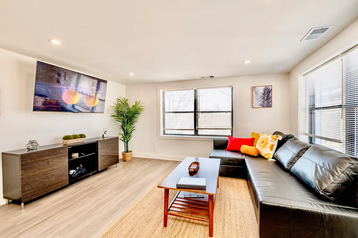 This image showcases a black leather sofa adorned with colorful cushions, a large TV mounted on the wall, a decorative plant, and a modern coffee table arranged in a cozy living room. The space looks comfortable, inviting, and designed for relaxation and entertainment.